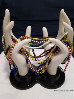 A white plastic jewelry holder, a hand holding beaded necklaces in different color patterns.