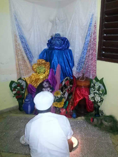 Image of Alo Ocan Roberto Gonzalez sitting with his back facing us, in front of him is a religious altar with a which decorative dove. 